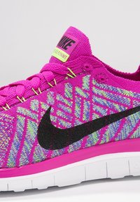 Růžová sportovní obuv vyrobená z texturovaného pleteného materiálu, se černým logem Nike, živými vzorovanými akcenty v modré a zelené barvě a bílou podrážkou.