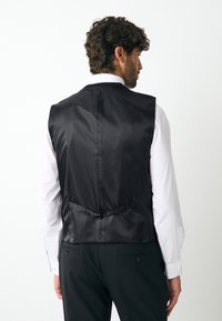 Gilet en satin noir à texture lisse, doté d'une boucle au dos et d'une coupe structurée, porté par-dessus une chemise blanche.