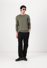 Pulover verde măsliniu cu guler rotund, manșete și tiv ribbate. Pantaloni negri și adidași. Ochelari de soare completează ținuta. Stil simplu, casual.