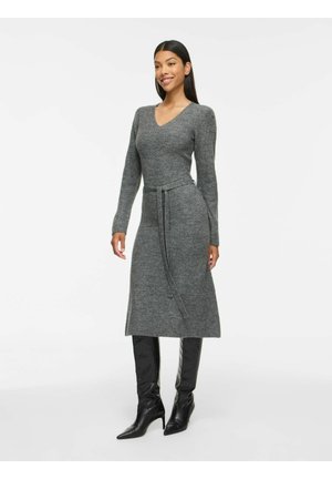 Femme portant une robe grise en tricot à manches longues avec une ceinture à la taille et des bottes noires montantes à talons, debout et souriante sur fond blanc.