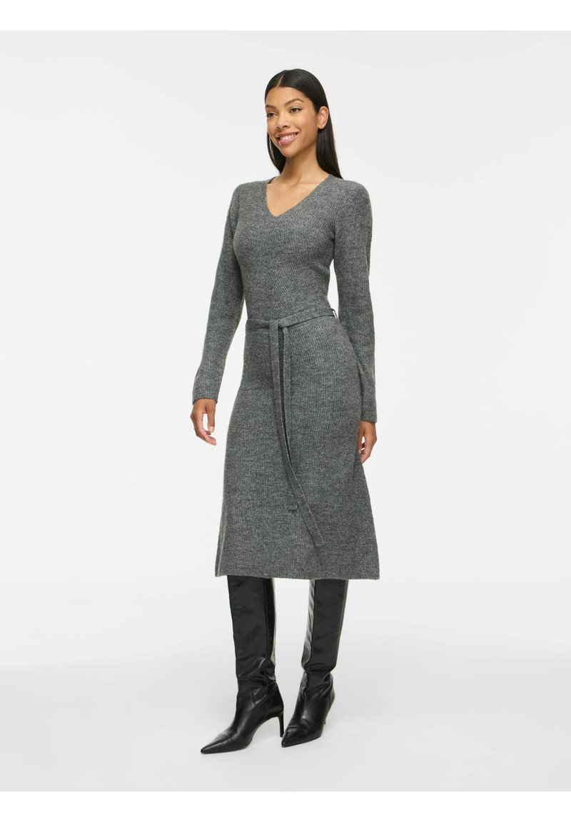 Femme portant une robe grise en tricot à manches longues avec une ceinture à la taille et des bottes noires montantes à talons, debout et souriante sur fond blanc.