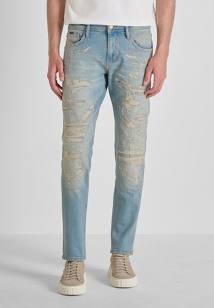 Lichtblauwe gescheurde spijkerbroek gedragen door een staande persoon, gecombineerd met beige sneakers en een wit shirt.