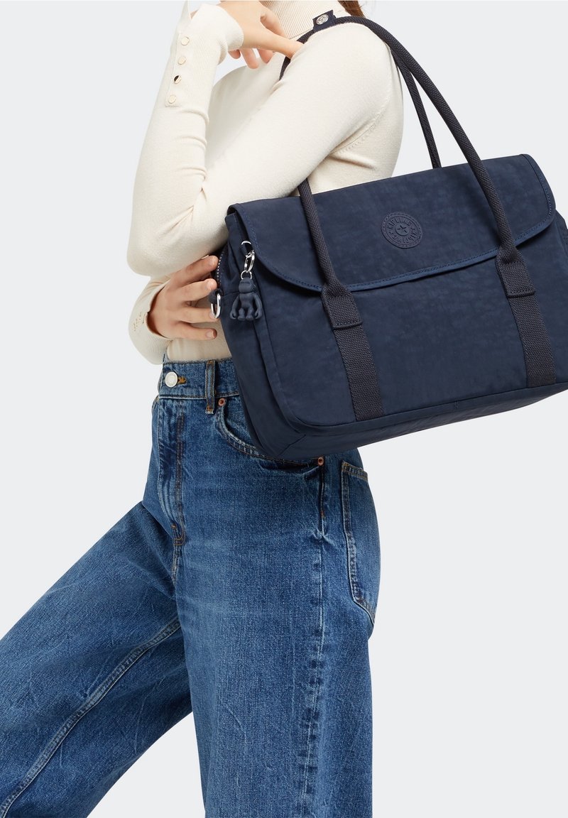 Femme portant un jean bleu et un pull crème porte un sac à main bleu marine avec de longues anses sur l'épaule.