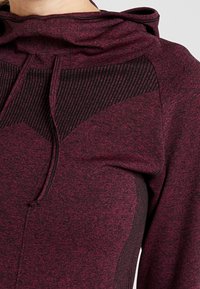 Burgundy hoodie i texturerat tyg med ett svartrandigt mönster på den övre bröstkorgen och dragsnöre i huvan.