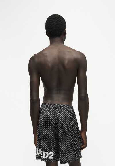 Homme à la peau foncée et aux cheveux courts bouclés, torse nu, portant un short noir à motifs avec le texte "DSQUARED2" dans le dos, debout devant un fond blanc uni.