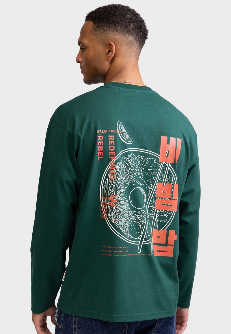Uomo con camicia verde a maniche lunghe con grafica rossa e bianca di una ciotola di noodles e bacchette, oltre a testo in inglese e coreano sul retro.