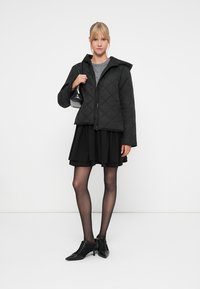 Črna prešita jakna s kapuco, v kombinaciji s sivo majico, slojevno črno krilo in črne hlačne nogavice do kolen. Dodatki vključujejo majhno torbico in čevlje z vezalkami.