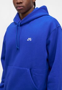 Άτομο που φοράει μπλε φούτερ Nike SB με κορδόνια και μπροστινή τσέπη, εμφανίζοντας τον άνω κορμό και μέρος του προσώπου.