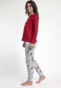 Mujer vestida con una blusa de manga larga roja y pantalones de pijama gris claro con estampados de cartas de juego y relojes, de pie descalza contra un fondo blanco.