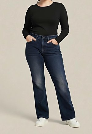 Personne portant un jean droit bleu foncé, un haut à manches longues noir, des baskets blanches, debout les mains dans les poches devant un fond uni.