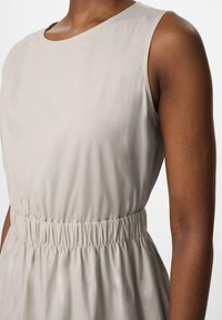Robe sans manches beige clair avec un col rond, une taille élastique et une texture de tissu lisse et légèrement brillante.