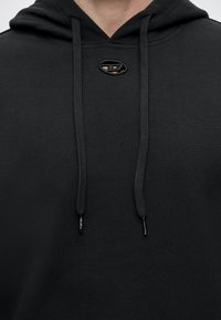 Gros plan d'un sweat à capuche noir avec des cordons et un petit logo ovale près du col.