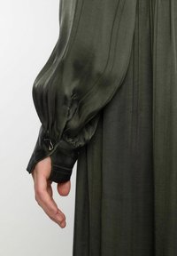 Robe olive verte avec une longue manche plissée et un poignet ajusté, dotée de détails en métal argenté, fabriquée à partir d'un tissu lisse et brillant.