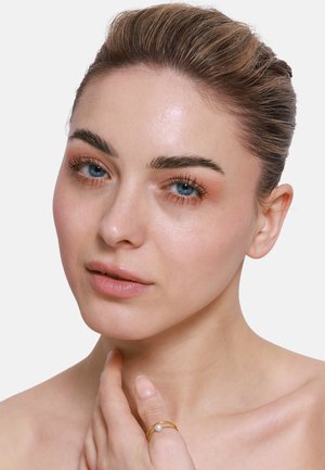Nahaufnahme einer Person mit glatter, heller Haut, blauen Augen und einem subtilen Glanz. Sie trägt einen zarten Goldring mit einem Perlenakzent.