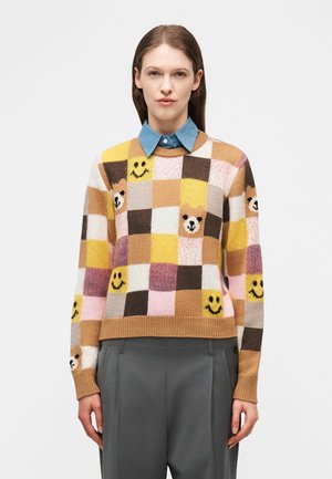 Frau trägt einen mehrfarbigen karierten Pullover mit Bärengesichtern und Smiley-Mustern, kombiniert mit grauer Hose und blauem Hemd mit Kragen.