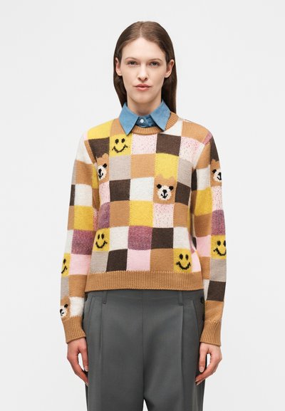 Femme portant un pull à carreaux multicolores orné de visages d'ours et de motifs souriants, associé à un pantalon gris et une chemise bleue à col.