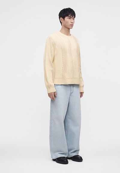 Homme portant un pull en maille crème, un jean large bleu clair et des chaussures noires, debout devant un fond clair uni.