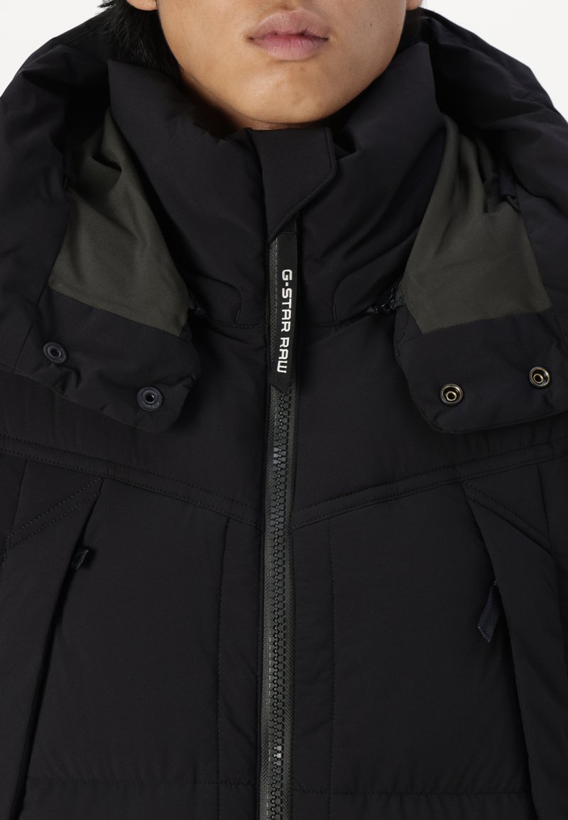 Chaqueta aislante negra con cuello alto, cierre de cremallera y marca G-Star Raw. Presenta detalles de botones y costuras geométricas.