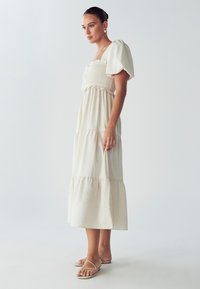 Šviesiai smėlinis maksi suknelė su pūstomis rankovėmis, smulkinta liemenėlė, sluoksniuota sijonas ir delikatūs volanai. Dėvima su plokščia, su dirželiais šlepečių.