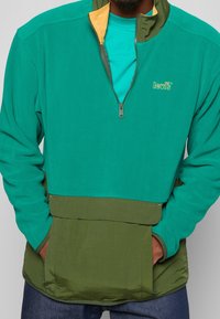 Grøn og blågrøn pulloverjakke med halv lynlås, kontrasterende sennepsfarvet krave og en stor lomme foran. Blødt tekstureret stof.