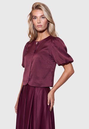 Blonde Frau trägt eine glänzende dunkelviolette kurzärmelige Bluse mit Puffärmeln und passenden plissierten Rock, steht vor einem einfarbigen Hintergrund.