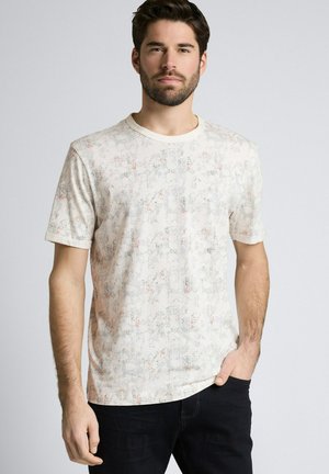 Mężczyzna z ciemnymi włosami i brodą, ubrany w jasnokolorowy, krótki rękaw koszulkę w wzory oraz czarne spodnie, stojący z jedną ręką w kieszeni.