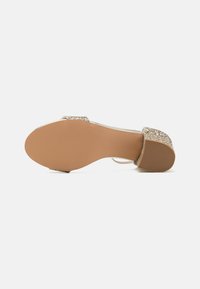 Högklackad sandal med en glittrig guldrem, genomskinligt material och en slät beige sula. Yttersulan har subtila ribbor för grepp.