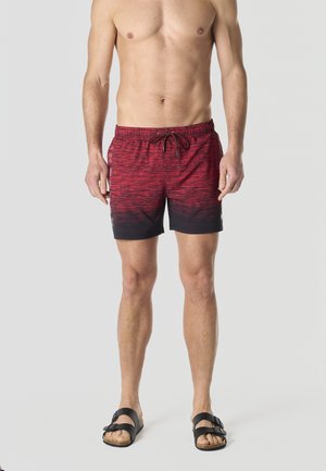 Hombre sin camiseta vistiendo pantalones de baño en degradado rojo y negro con texto lateral y sandalias negras con hebilla, de pie contra un fondo liso.
