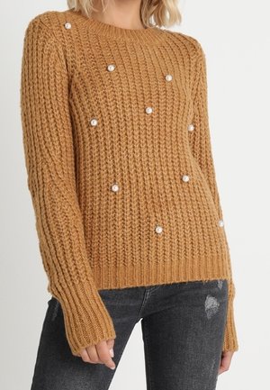 Vrouw draagt een beige grofgebreide trui met parelversieringen, gecombineerd met zwarte distressed jeans, tegen een effen achtergrond.