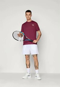 Bordo sporta krekls, balti šorti, tur rokās tenisa nūju ar melnu un zilu rāmi. Gluda auduma, minimālistisks dizains un redzami tetovējumi.