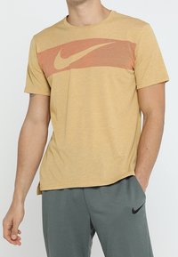 Hombre vestido con una camiseta Nike de color mostaza con una franja horizontal naranja y el logo de swoosh, combinada con pantalones de chándal Nike grises con el logo de swoosh en negro.