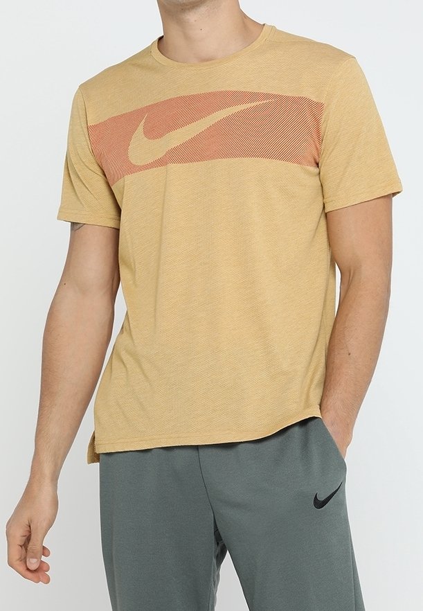 Hombre vestido con una camiseta Nike de color mostaza con una franja horizontal naranja y el logo de swoosh, combinada con pantalones de chándal Nike grises con el logo de swoosh en negro.