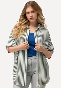 Camicia a righe con bottoni color beige e blu navy, maniche arrotolate e dettagli rossi; indossata sopra una maglietta blu e pantaloni grigi chiari.