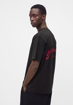 Ung mand med kort hår iført en løs sort T-shirt med rød tekst på ryggen, stående sidevendt med hænderne i lommerne.