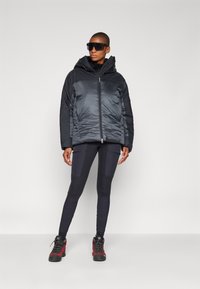 Giubbotto imbottito grigio scuro con cappuccio, dotato di zip frontale e design a righe. Abbinato a leggins neri e stivali da trekking rossi.
