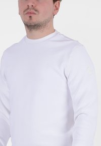 Camicia bianca a maniche lunghe con scollatura rotonda e tessuto a costine. Presenta un logo discreto sulla manica sinistra. Design semplice e pulito.