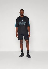 Černé sportovní tričko s modrými proužky a nápisem „ADIDAS SPORTSWEAR“; doplněné černými šortkami a černými sportovními botami.