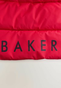 Chaqueta de abrigo roja con una textura gruesa y acolchada. Presenta grandes letras negras que dicen "BAKER" en la parte del pecho. Acabado de material suave.