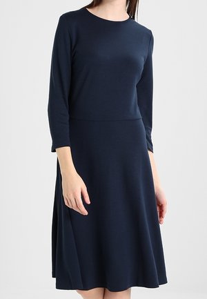 Nainen, joka pukeutunut tummansiniseen, pitkähihaiseen, polvipituiseen mekkoon, jossa on istuva vyötärö ja pyöreä pääntie vaaleaa yksiväristä taustaa vasten.