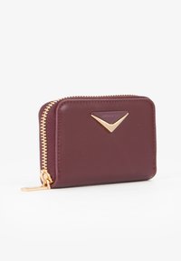 Portafoglio in pelle bordeaux compatto con zip dorata ed emblema dorato triangolare sul davanti, posizionato su uno sfondo bianco.