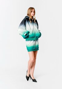 Chaqueta con capucha de diseño ombré en negro, blanco y verde azulado, combinada con una falda a juego. La modelo lleva zapatos de tacón negros.