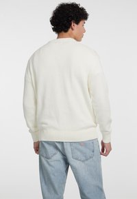 Cremefarvet strikket sweater med lange ærmer, ribbet kant og en afslappet pasform. Bæres med lyseblå denimjeans med et trekantet logo.