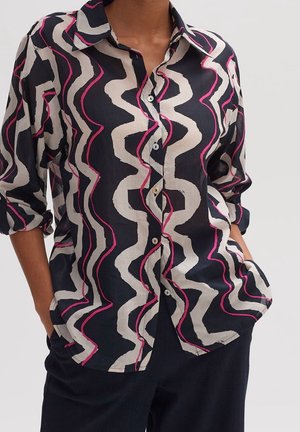 Femme portant une chemise noire boutonnée avec des rayures ondulées beige et rose, les mains glissées dans les poches d'un pantalon bleu marine.