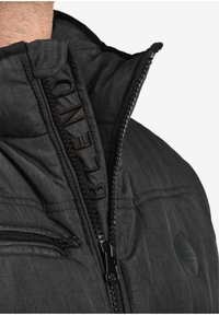 Schwarze Steppjacke mit hohem Kragen, strukturiertem Stoff, schwarzen Reißverschlüssen und einem bestickten Logo auf der Vorderseite. Verfügt über eine Reißverschlusstasche.