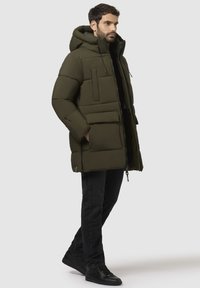 Olivengrøn pufferjakke med hætte, der har lynlås foran, lommeapplikationer og polstret design. Sammen med sorte jeans og sneakers.