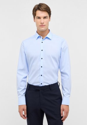 Chemise à manches longues bleu clair avec un col pointu, des boutons noirs et une texture lisse, portée avec un pantalon sombre.