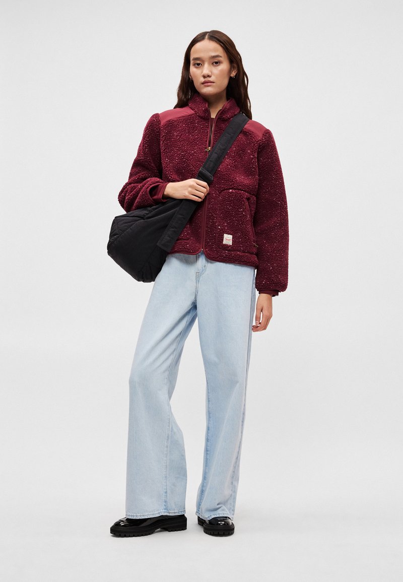 Burgundy fleece jacket with a zip, two front pockets, and black accents. Paired with light blue wide-leg jeans and black platform boots.