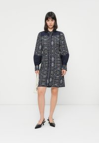 Navy patterned dress with long, puffed sleeves and a button closure. Features a contrasting collar and intricate floral print, paired with black heels.