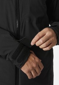 Veste noire imperméable avec des poignets ajustables en Velcro. Texture lisse avec une fermeture éclair à l'avant et une coupe moulante.