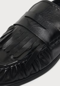 Μαύρο δερμάτινο loafer με διακοσμητικό κρόσσι και ραμμένες λεπτομέρειες στην επάνω πλευρά και γύρω από την στρογγυλεμένη περιοχή των δαχτύλων.